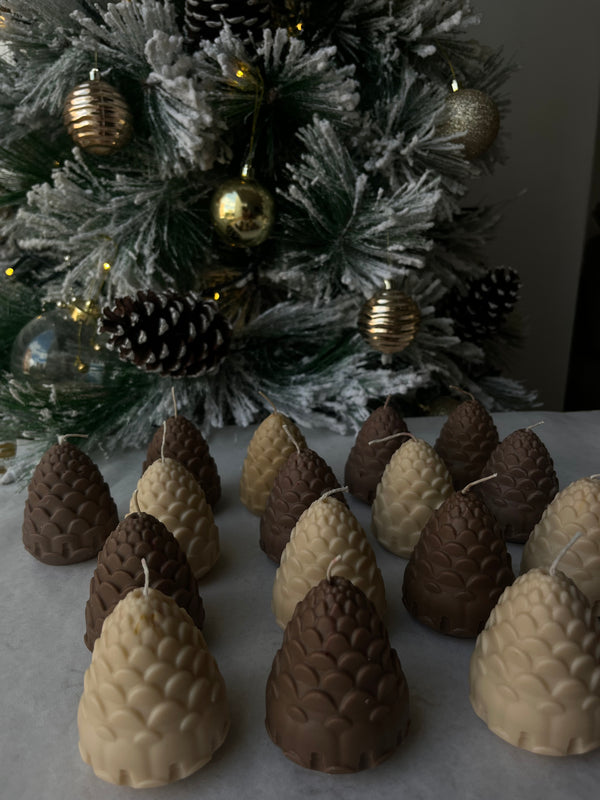 Piñas de Navidad de La Marie en cera vegetal, en crema, beige y chocolate, sobre una mesa con un árbol nevado