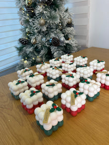 Cubos navideños de La Marie en cera vegetal, con acebo y etiqueta dorada, sobre una mesa de madera frente a un árbol nevado