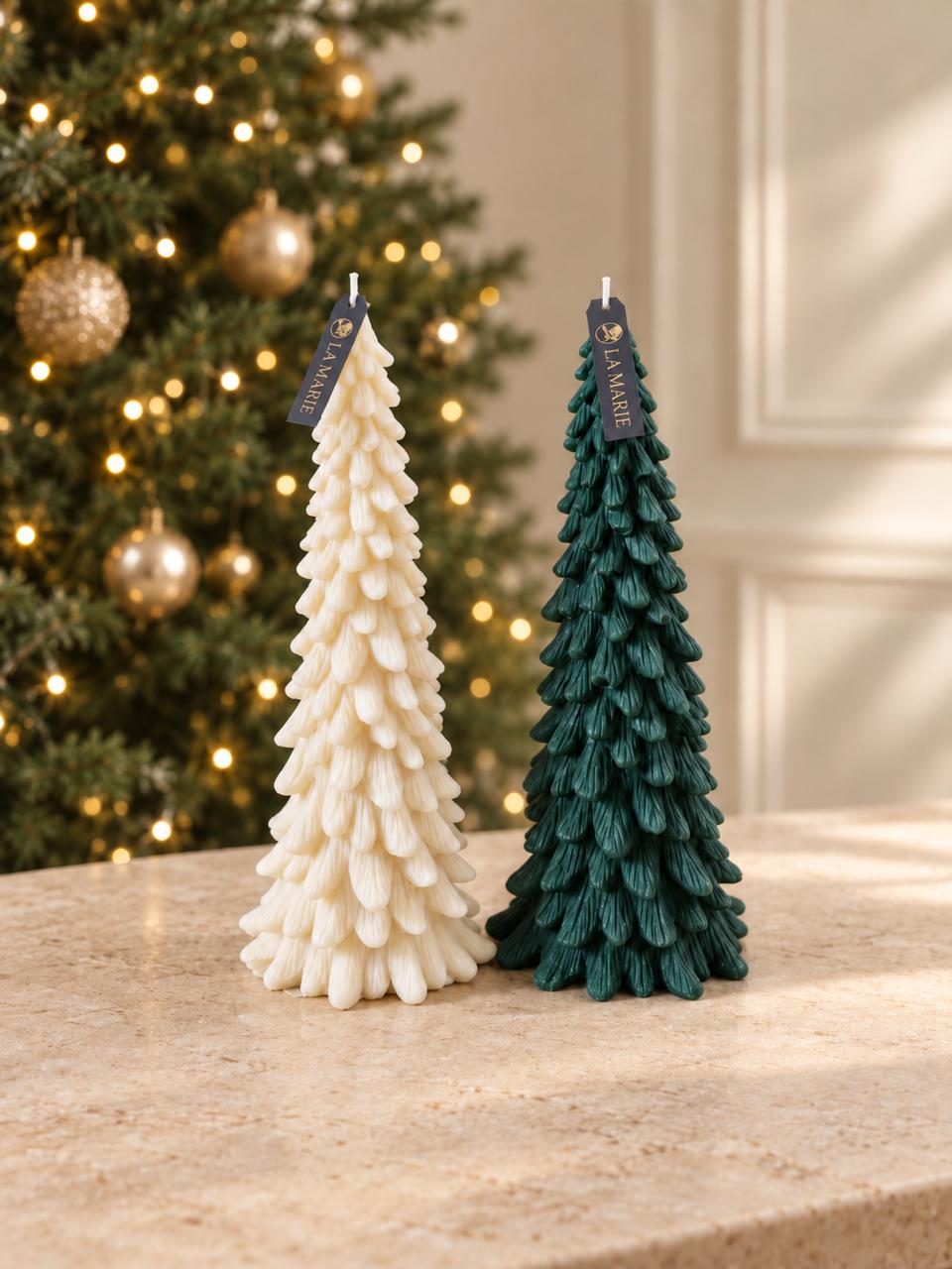 Dos pinos de La Marie, uno crema y uno verde, sobre una mesa de mármol frente a un árbol de Navidad con luces