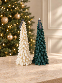 Dos pinos de La Marie, uno crema y uno verde, sobre una mesa de mármol frente a un árbol de Navidad con luces